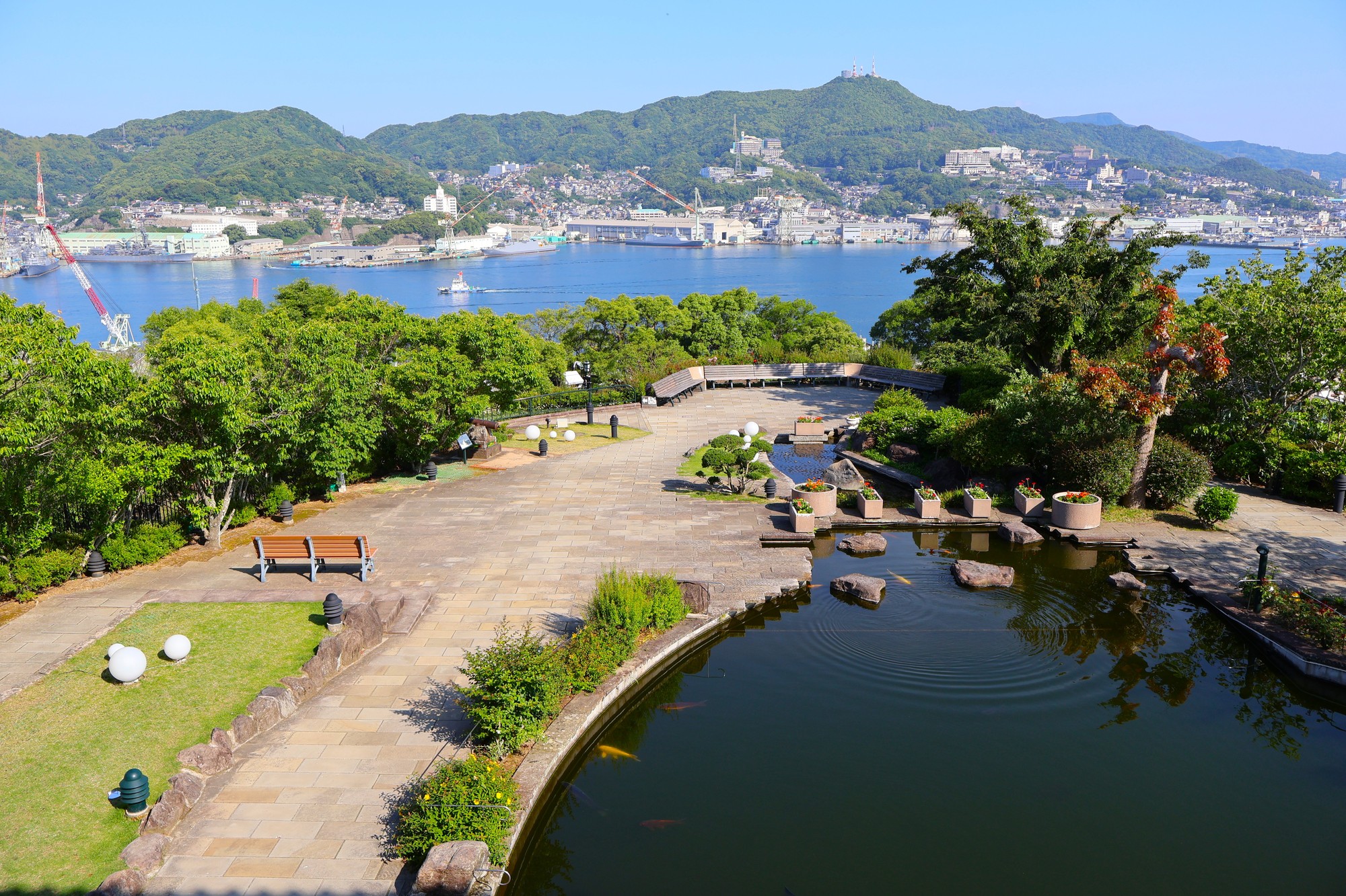 グラバー園の高い位置から見下ろした長崎港。手前に池と石畳の園路、対岸に造船所のクレーンが並ぶ